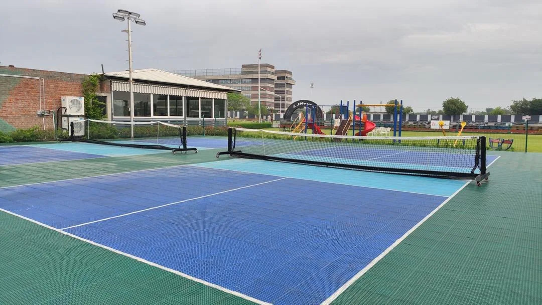 Outdoor pickleball courts with nets, a building, and a playground in the background.