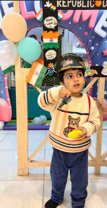A young child in a sweater and cap holds a toy, standing in front of a Republic Day decoration.