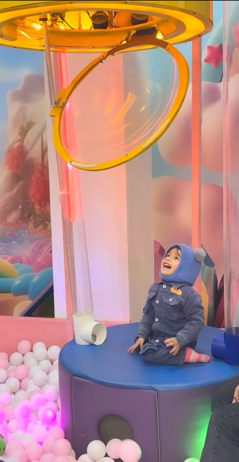 A young child in a blue hat and denim jacket sits on a blue platform, looking up at a yellow bubble machine in a ball pit.