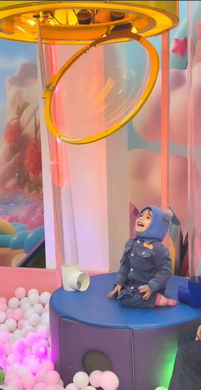 A young child in a blue hat and denim jacket sits on a blue platform, looking up at a yellow bubble machine in a ball pit.
