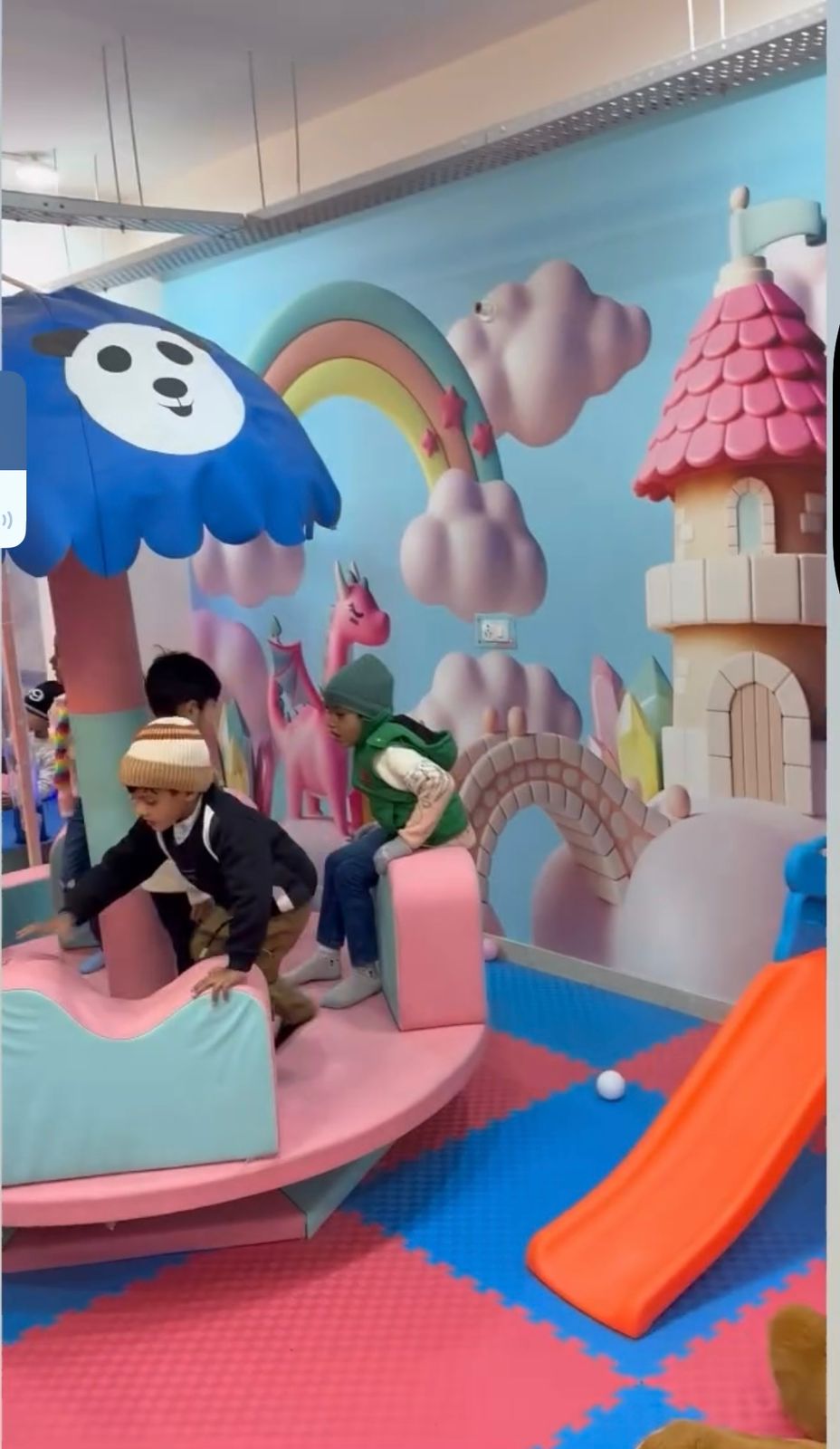 Children play in a colorful indoor playground with a carousel, slide, and fantasy-themed decorations.
