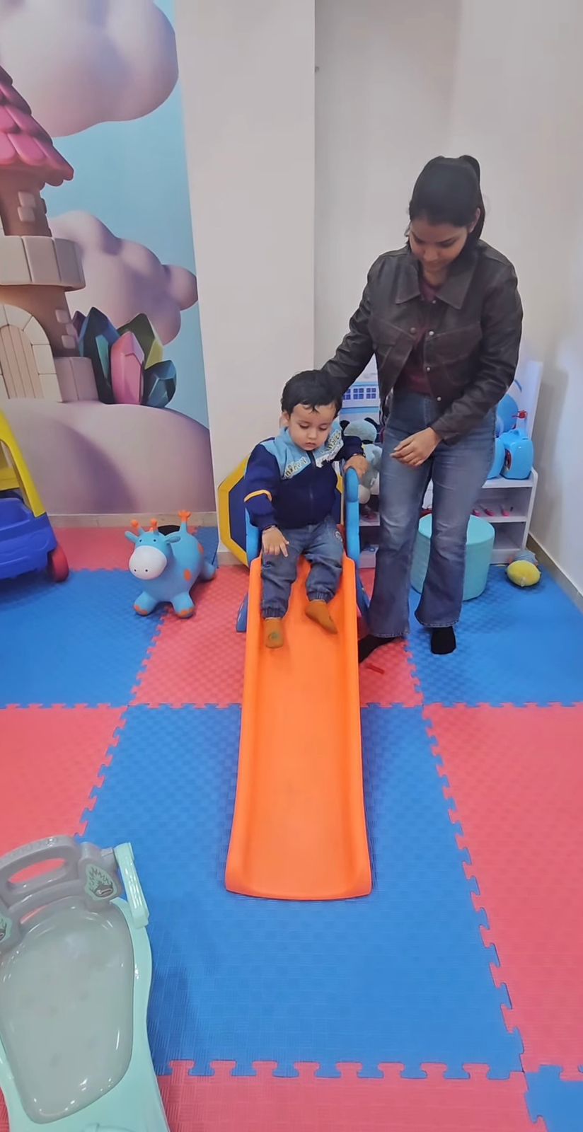 A child slides down a small orange slide while a woman supervises in a brightly colored playroom.