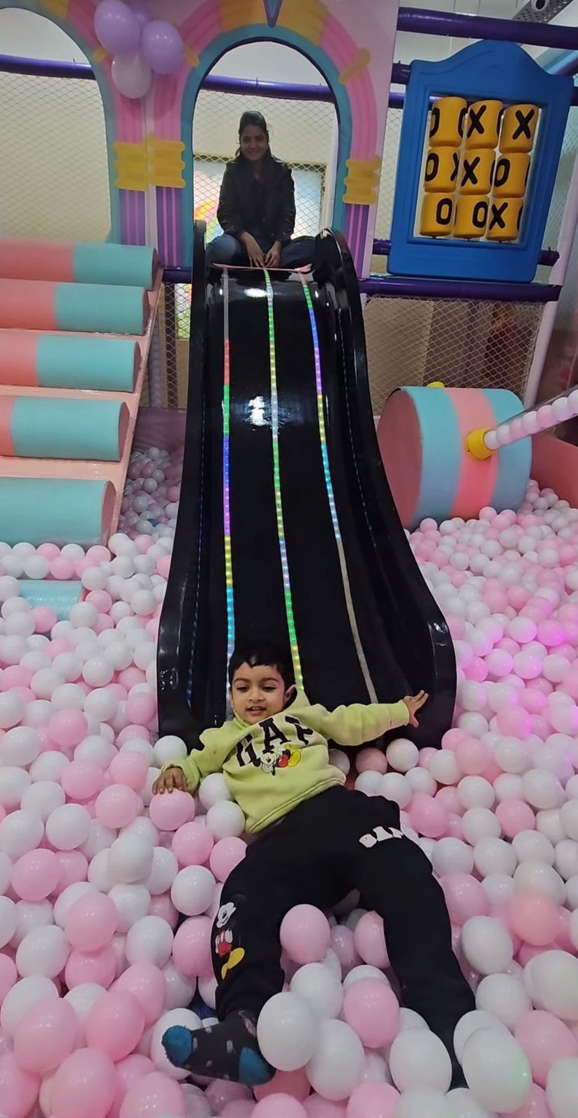 Child sliding down a colorful slide into a pink ball pit with another person at the top.
