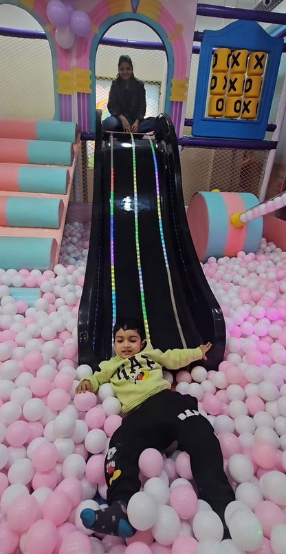 Child sliding down a colorful slide into a pink ball pit with another person at the top.
