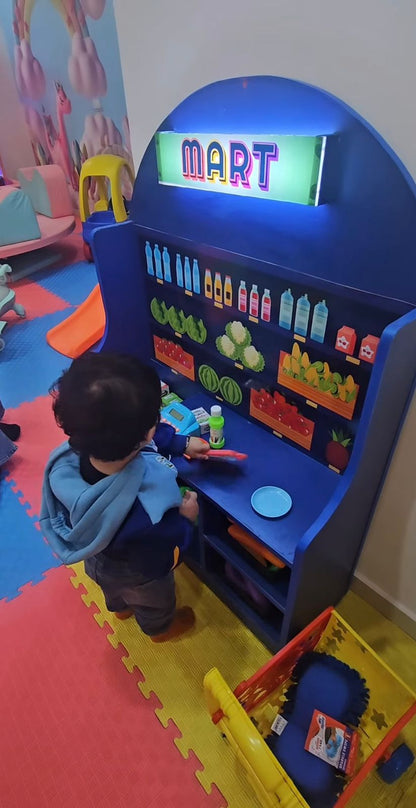 A young child plays at a toy grocery store with a "MART" sign. The child is interacting with the play food and cash register.