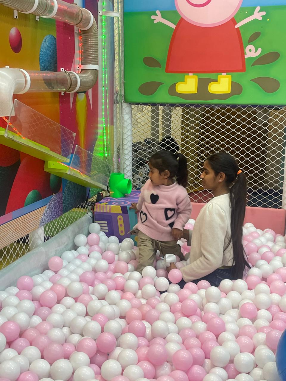 Two young children play in a ball pit with pink and white balls.