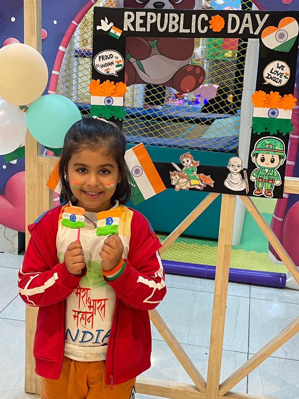 A young girl smiles, holding Indian flags, celebrating Republic Day. She has face paint and wears patriotic attire.