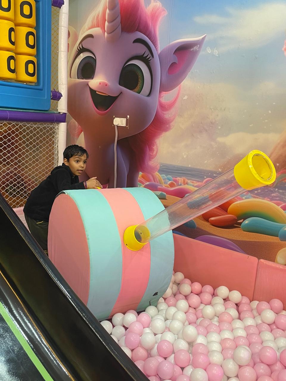 A young boy plays in a ball pit with a large unicorn mural in the background.