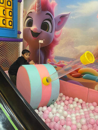 A young boy plays in a ball pit with a large unicorn mural in the background.