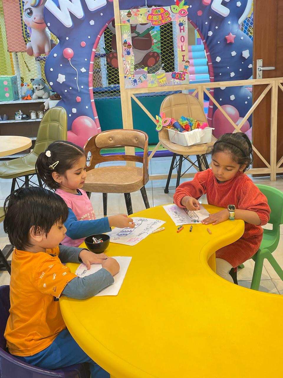 Three young children are drawing and coloring at a bright yellow table in a colorful indoor play area.