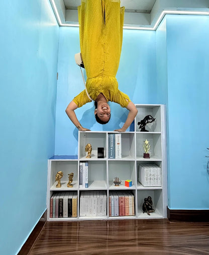 A young woman in a yellow dress is upside down, balancing on her hands in front of a bookshelf.