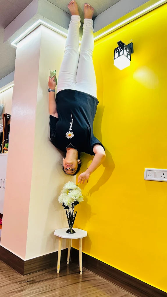 A young woman does a handstand against a wall, holding a green object. She wears a black t-shirt and white pants.