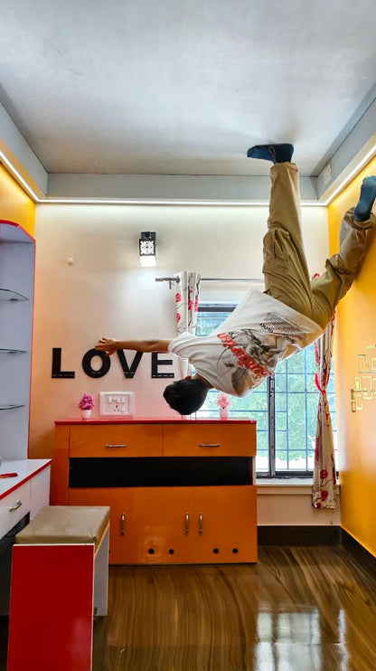 A young person does a handstand against a wall in a room with orange furniture and "LOVE" decor.