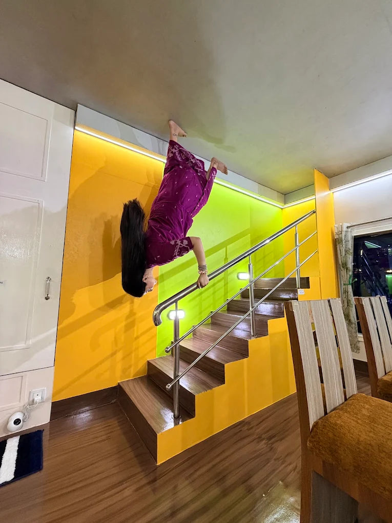 A person in a purple dress is upside down on a ceiling, appearing to walk down stairs. The room has bright yellow and green walls.