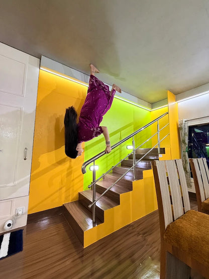 A person in a purple dress is upside down on a ceiling, appearing to walk down stairs. The room has bright yellow and green walls.