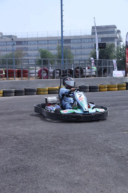 A person in a helmet drives a go-kart on a race track, with a building in the background.