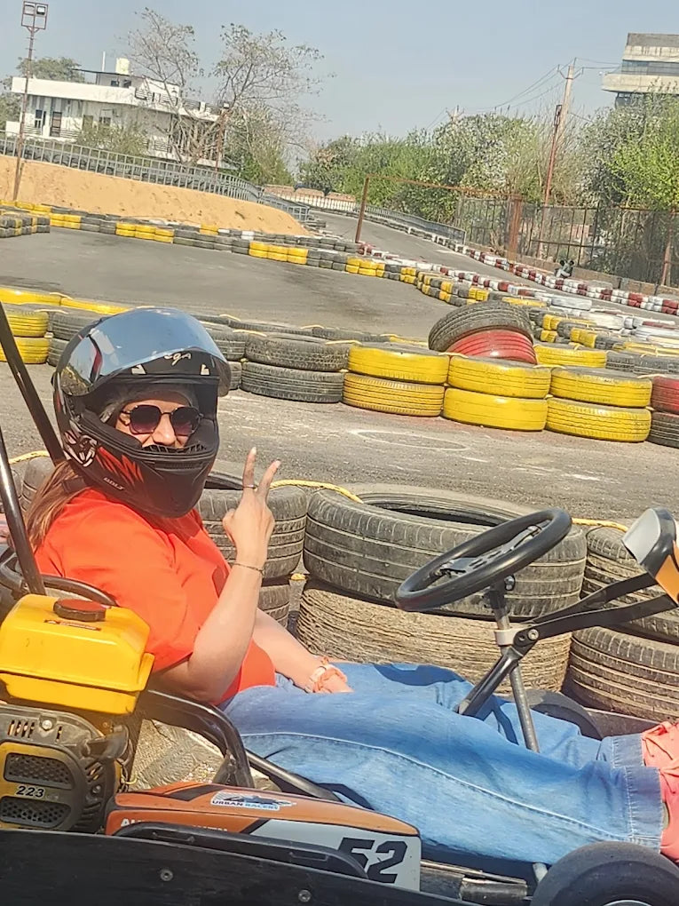 A young woman in a go-kart helmet and orange shirt makes a peace sign while sitting in a go-kart on a track.