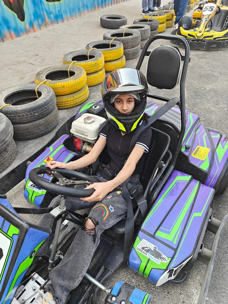 A young person in a go-kart helmet sits in a go-kart, ready to race. Tires are stacked in the background.