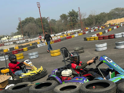 Two children race go-karts on a track, with a man walking in the background.