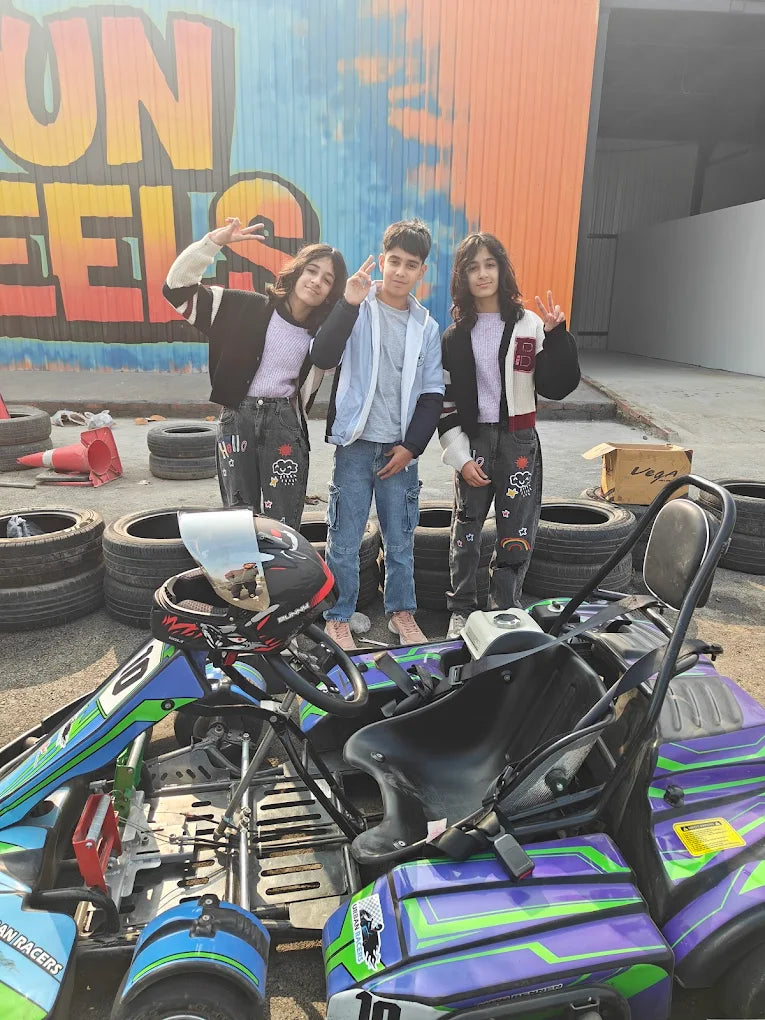 Three children pose in front of a go-kart with a colorful mural in the background.