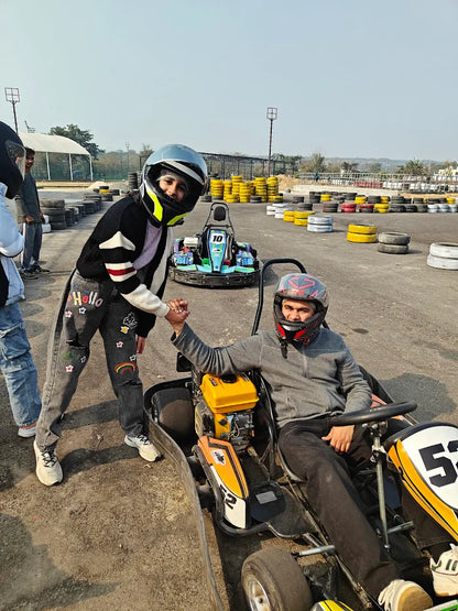 Two young people in helmets shake hands at a go-kart track, one in a go-kart and the other standing.