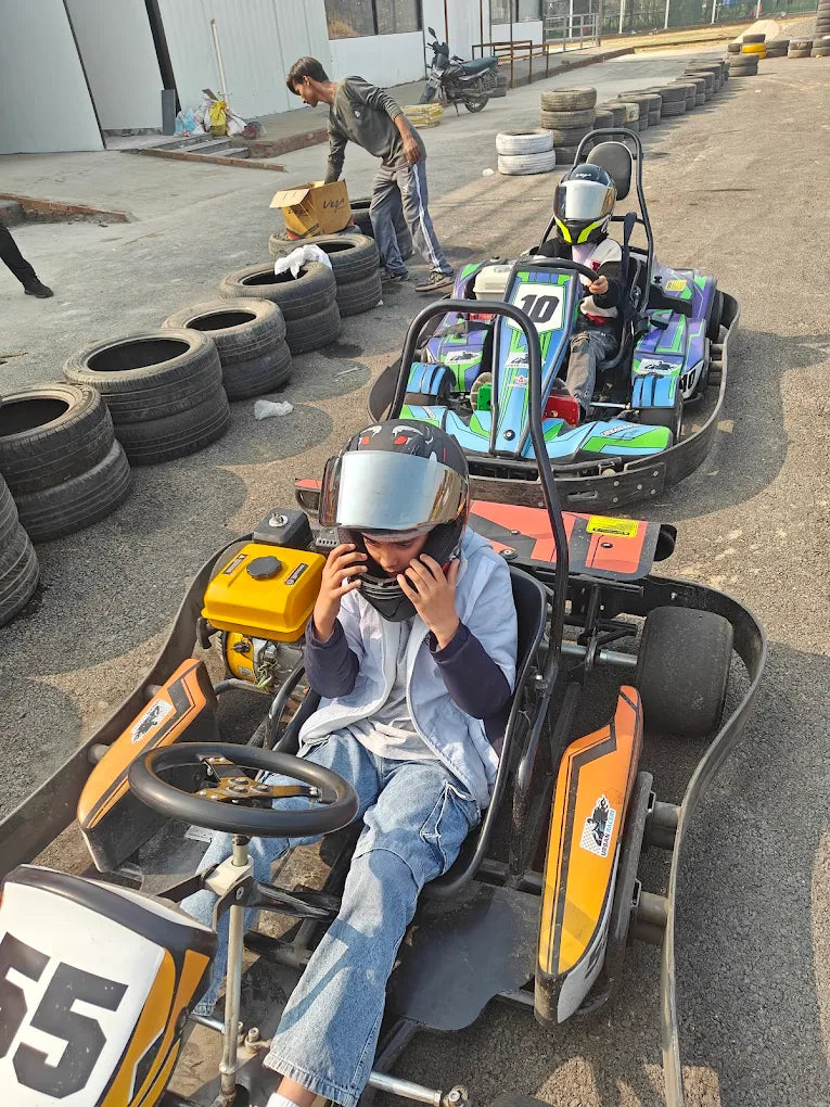 Two young children in go-karts on a track, one adjusting their helmet.