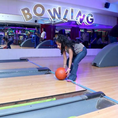A young girl prepares to bowl an orange ball at a bowling alley, with a "BOWLING" sign visible in the background.