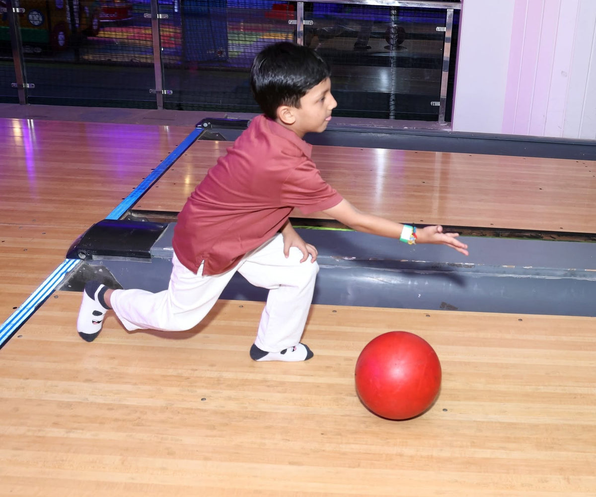 A young boy in a maroon shirt and white pants bowls a red ball down a wooden lane.