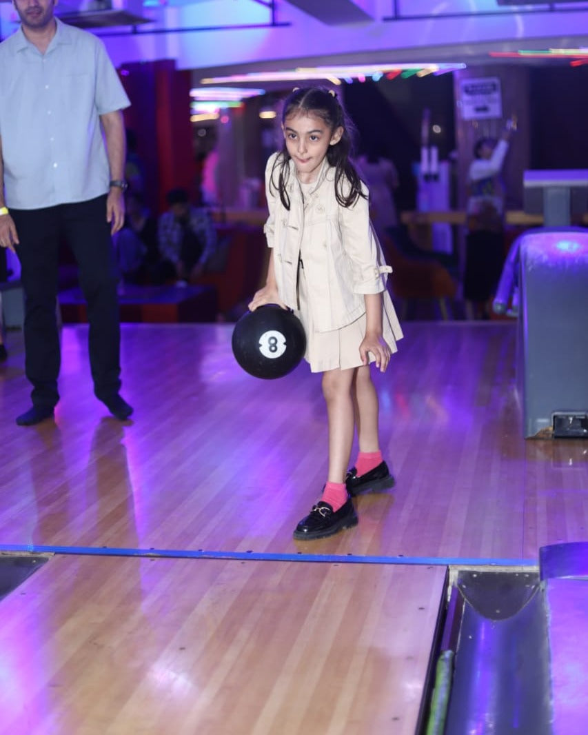 A young girl in a tan outfit holds a black bowling ball with the number 8 on it, ready to bowl.
