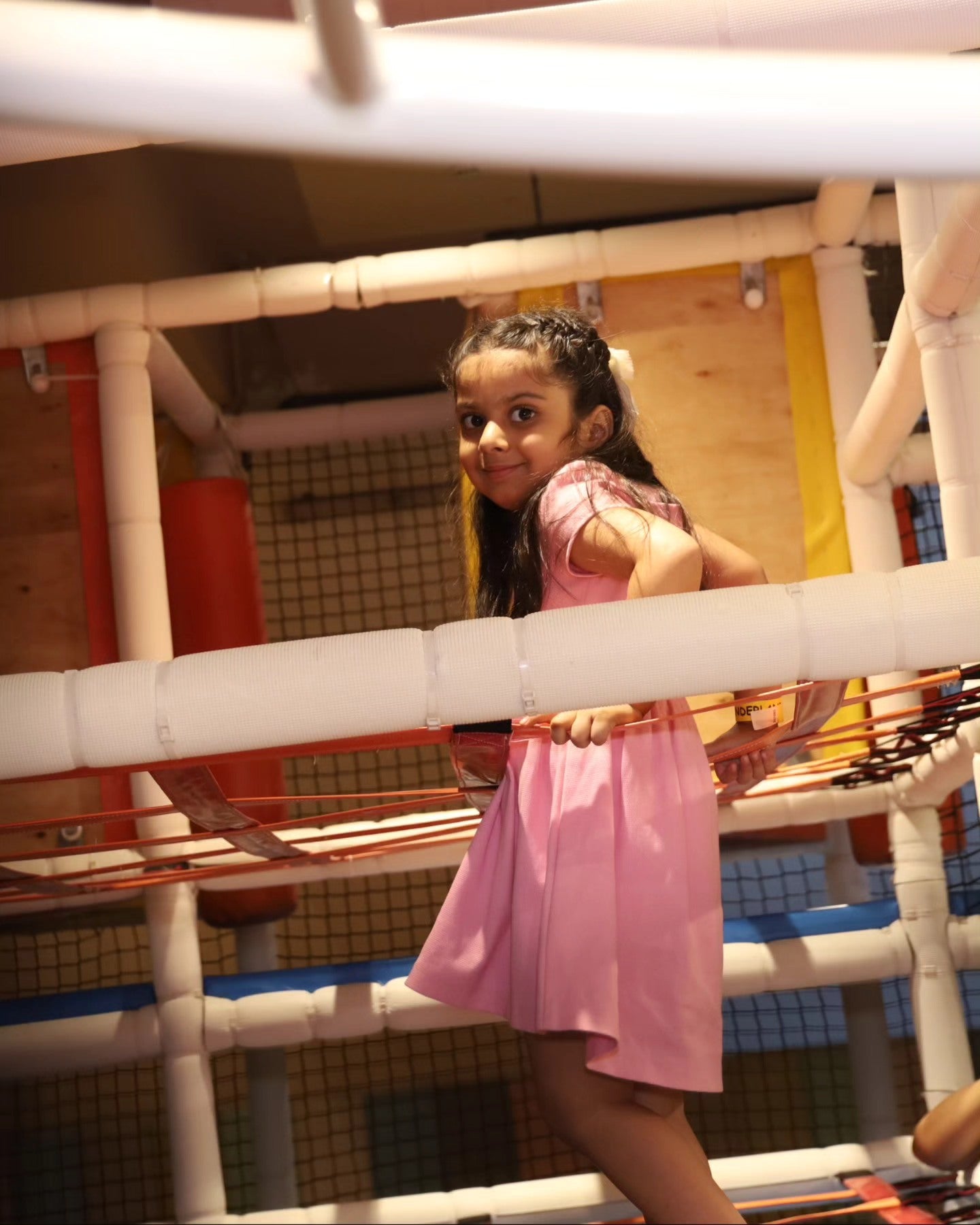 Young girl in a pink dress cautiously climbing vibrant rope barriers inside Walk to Wonderland Chhatarpur play area