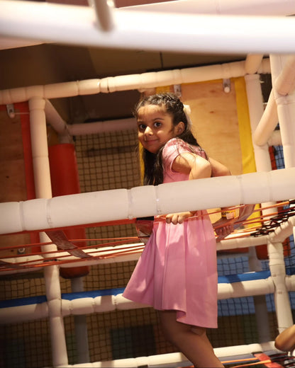 Young girl in a pink dress cautiously climbing vibrant rope barriers inside Walk to Wonderland Chhatarpur play area