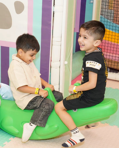 Two boys laughing and playing on a green animal-shaped rocker in Walk to Wonderland Indoor Play Area Chhatarpur
