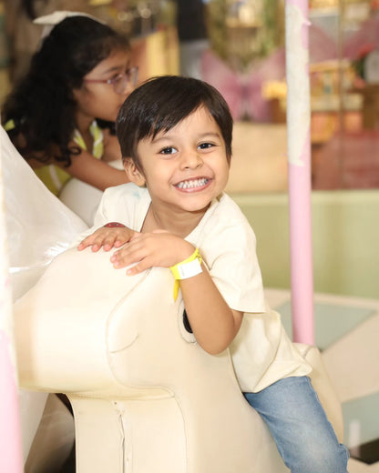 Smiling young boy wearing a yellow wristband playing on a white animal-shaped ride inside Play Area Chhatarpur