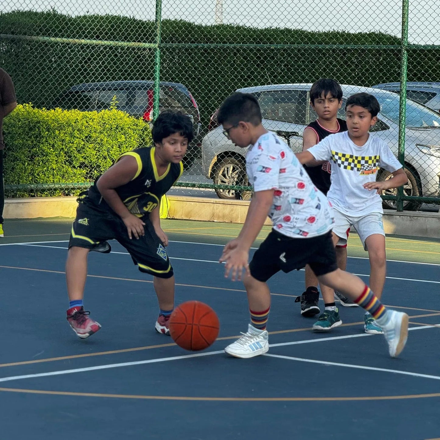 Children playing basketball on an outdoor court with cars and greenery in the background