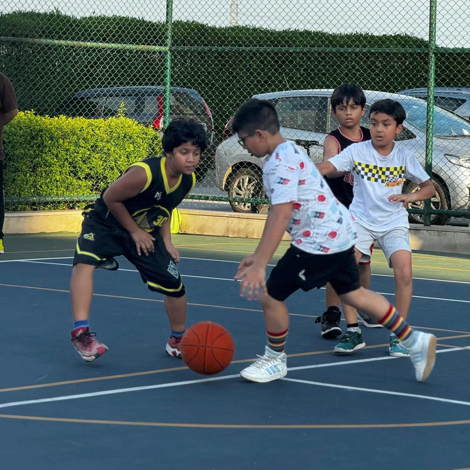 Children playing basketball on an outdoor court with cars and greenery in the background