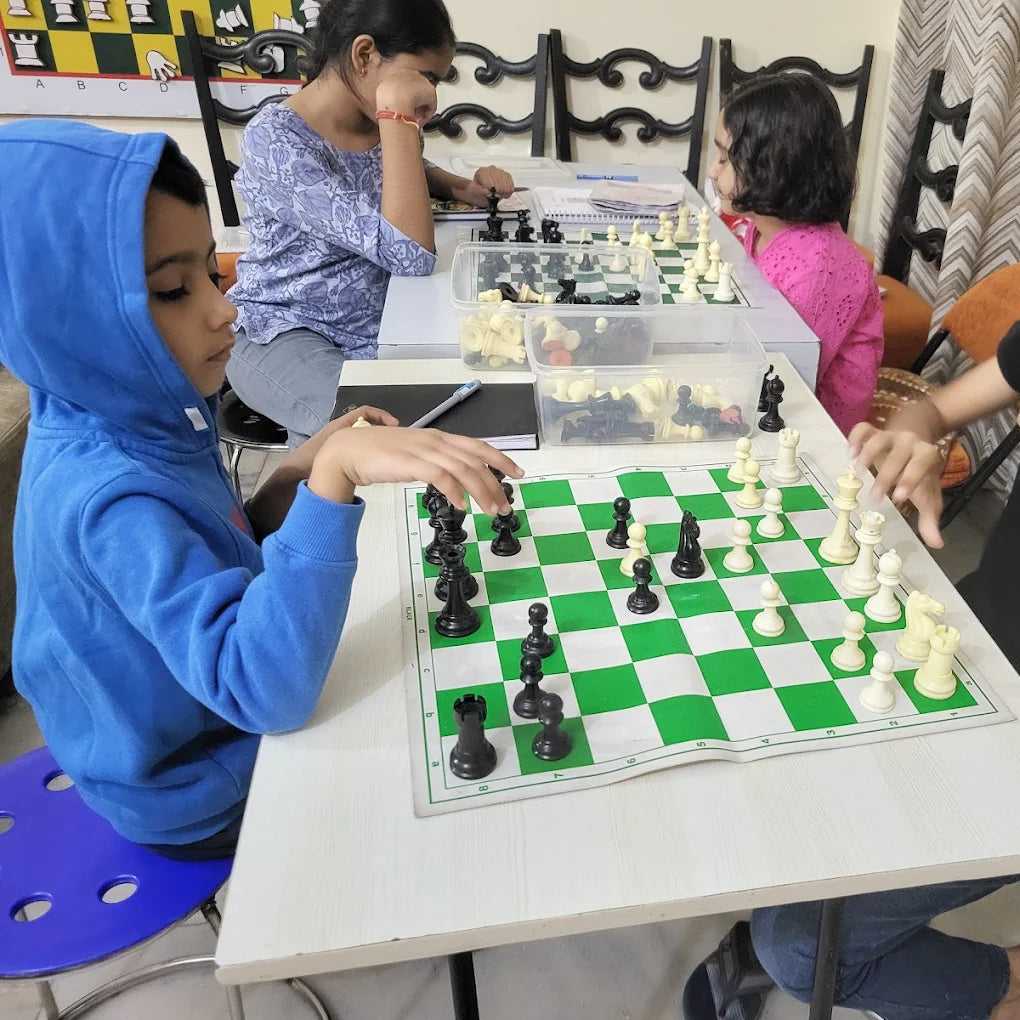Children playing chess at a table with other children in the background.