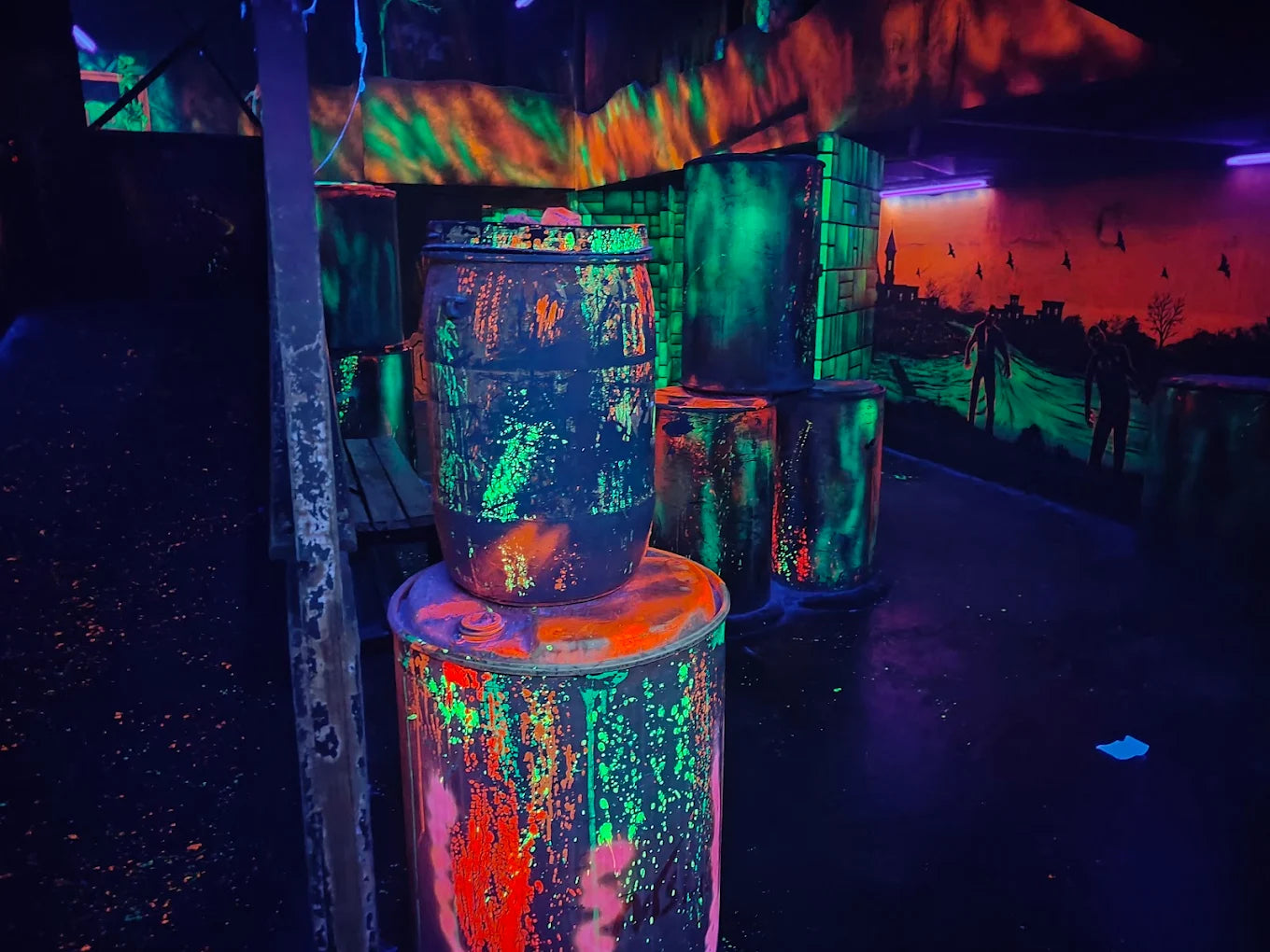 A neon-lit laser tag arena with barrels and zombie-themed murals.