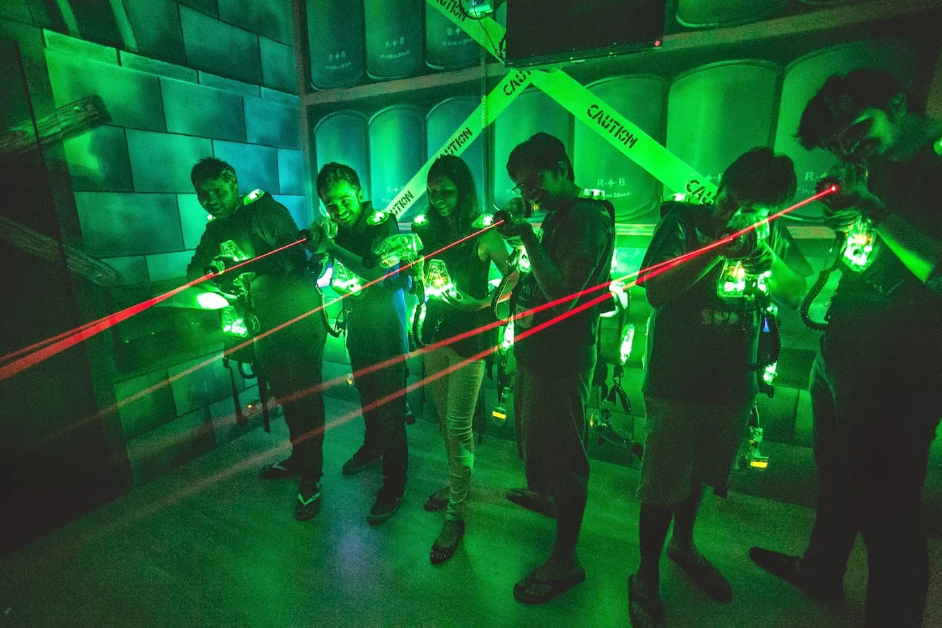 Young adults play laser tag in a dimly lit, green-lit arena, aiming their glowing phasers.