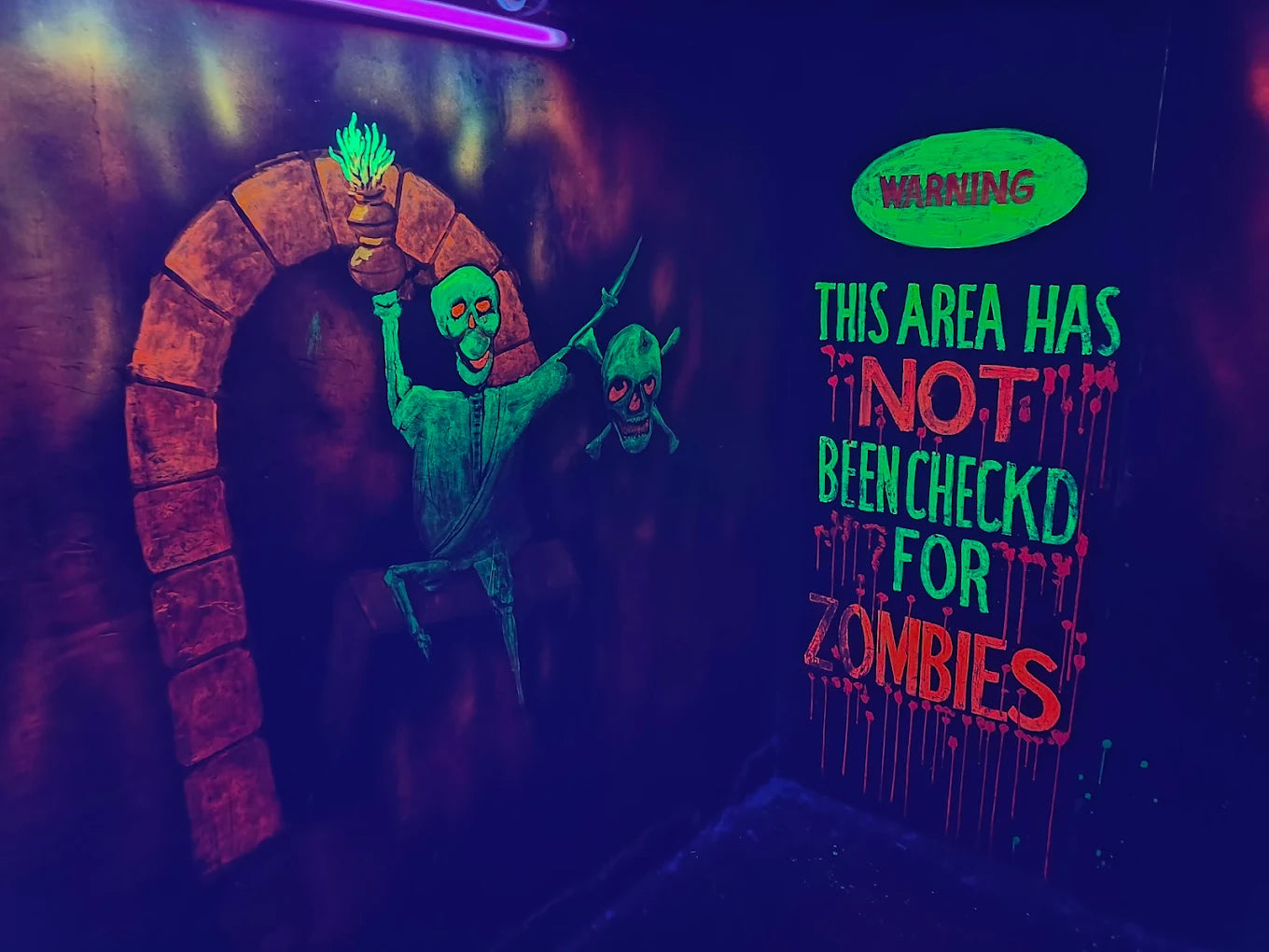 A neon mural in a dark room depicts skeletons and a warning sign about zombies.