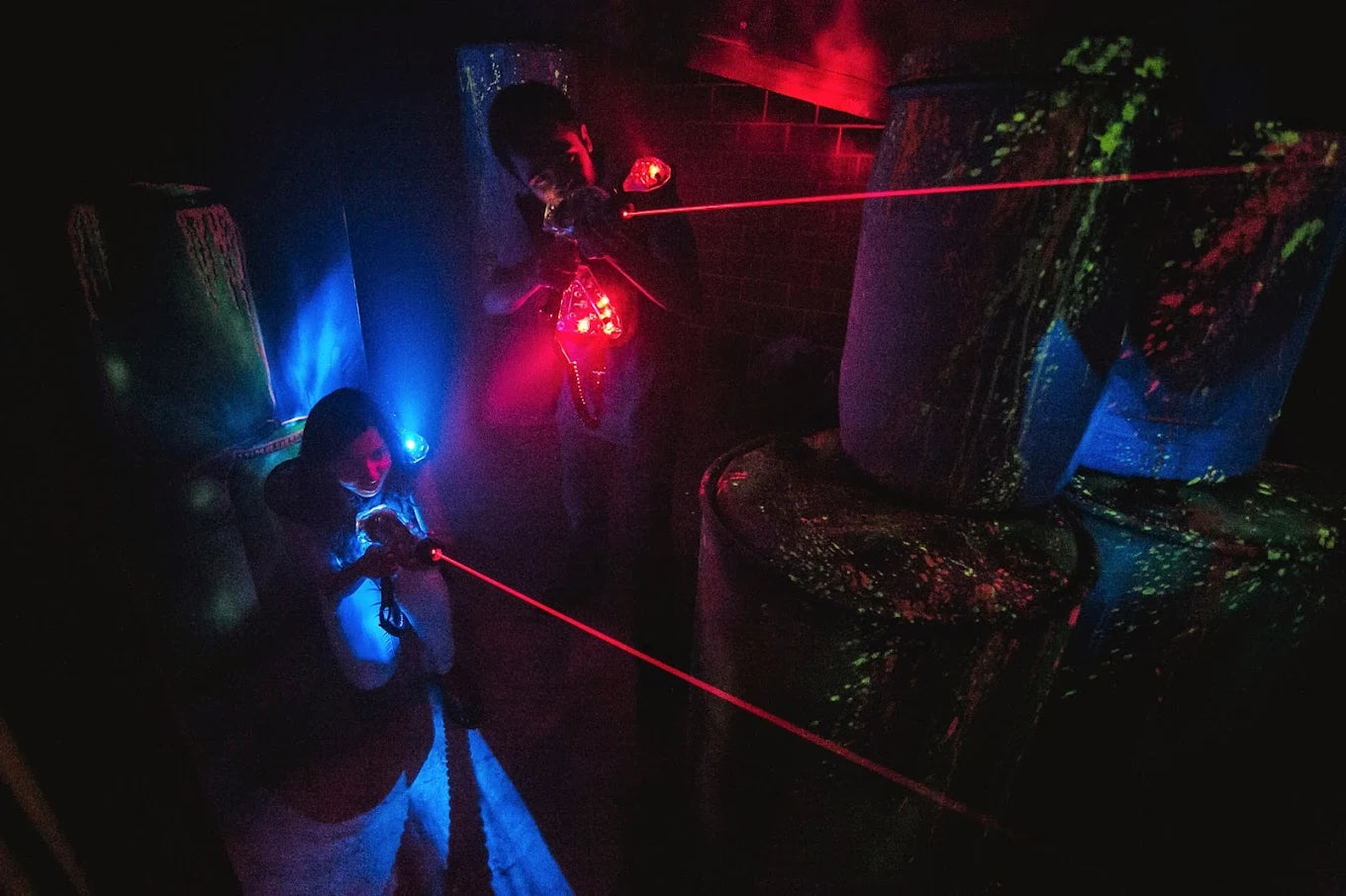 Young people play laser tag in a dark arena with glowing obstacles. Red and blue lights illuminate the scene as they aim their laser guns.