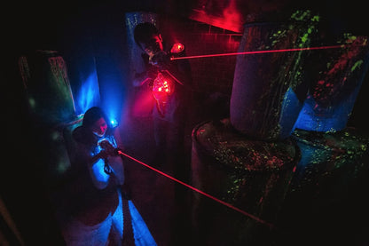 Young people play laser tag in a dark arena with glowing obstacles. Red and blue lights illuminate the scene as they aim their laser guns.