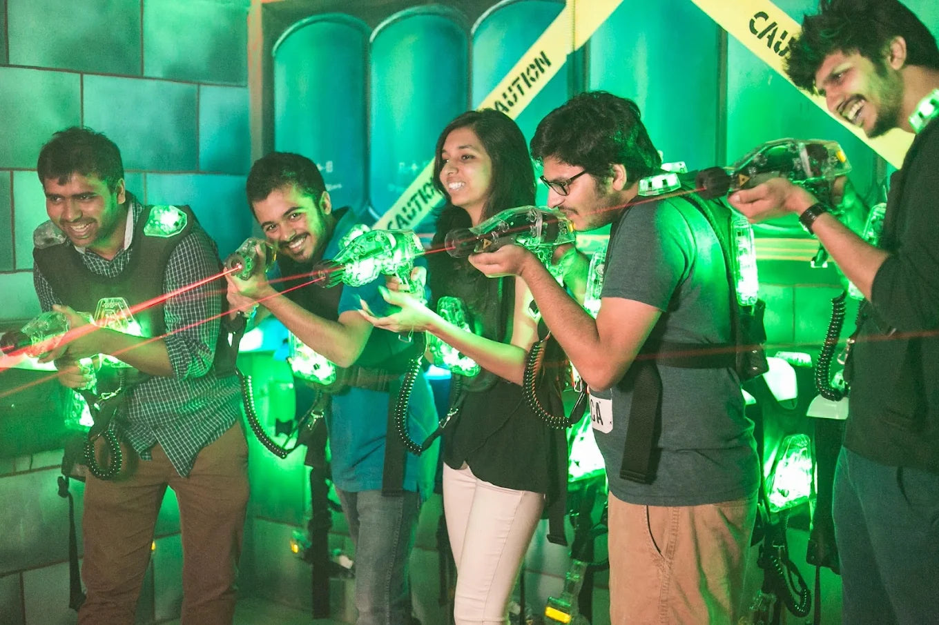Young adults play laser tag in a dimly lit, green-lit arena, holding glowing phasers.