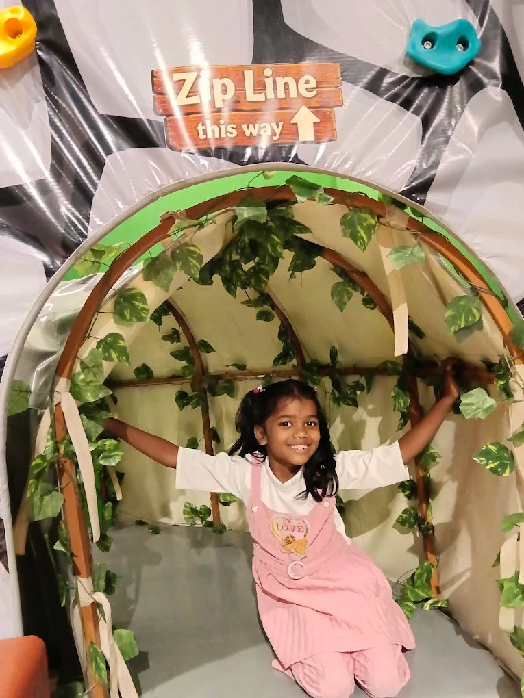 A young girl in a pink outfit smiles while holding onto a jungle-themed zip line entrance.