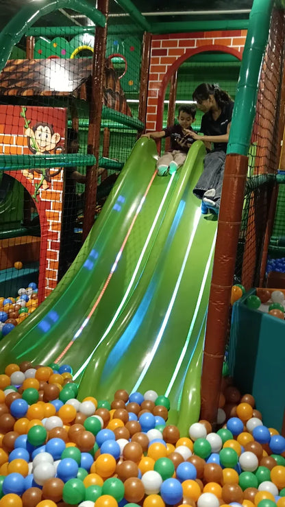 A child and an adult slide down a green playground slide into a ball pit.