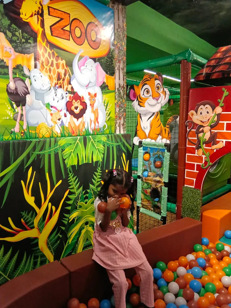 A young girl sits in a ball pit, playing with colorful balls, in a zoo-themed indoor playground.