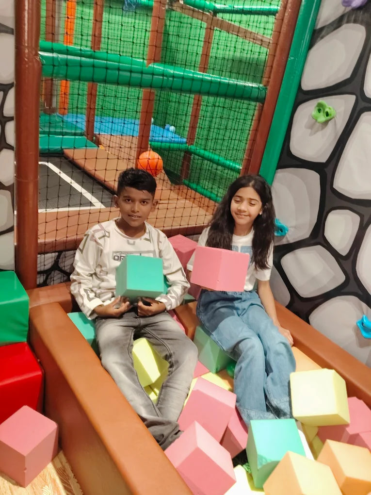 Two children sit in a ball pit with large foam cubes, surrounded by a colorful indoor playground.