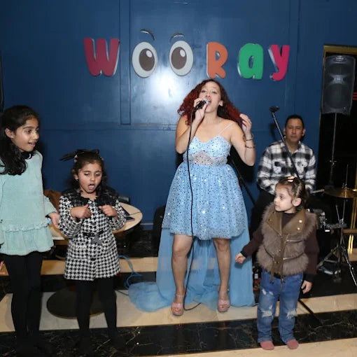 A young woman sings on stage while children watch, with a man in the background.