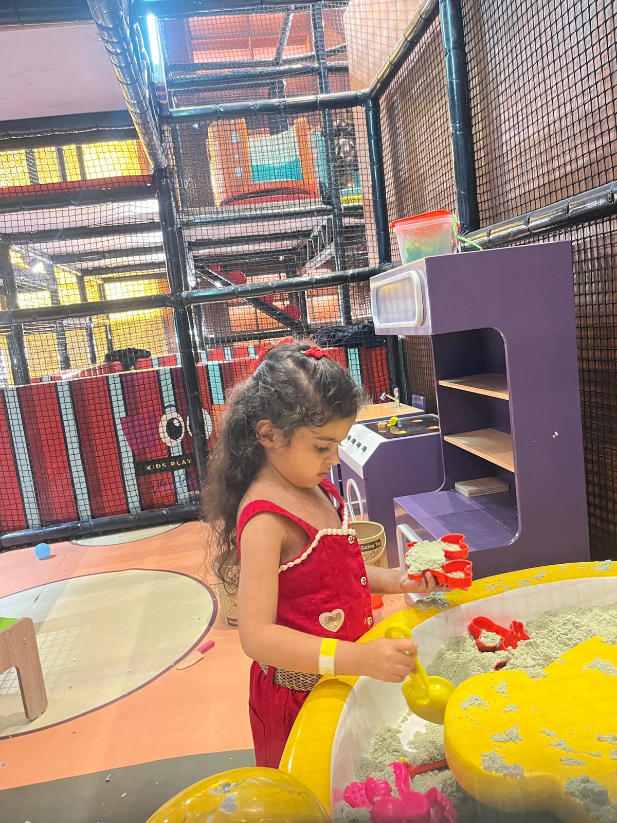 A young girl in a red outfit plays with sand toys in a colorful indoor play area.