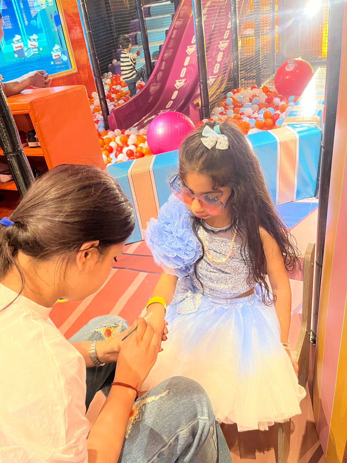 A young girl in a sparkly blue dress gets her arm painted by a woman in a play area with a ball pit.