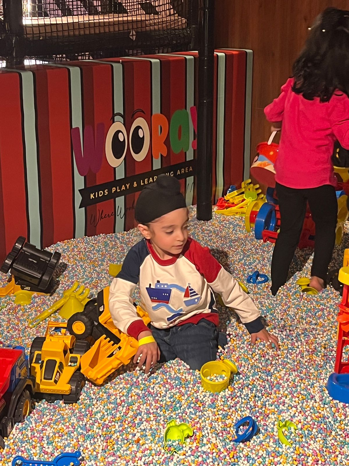 A young boy plays in a ball pit with toys, while a child stands nearby.